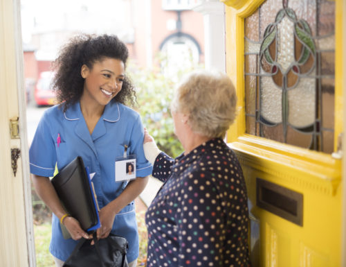 A female nurse or community care worker is at the front door of her senior female patient and saying hello. She is wearing a blue nurses tunic , and holding a medicines bag . She is wearing an ID badge with her profile photo already on it . The senior patient has her back to us at the front door . In the background a residential street can be seen defocussed .